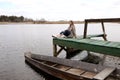 Young beautiful woman sits on the wooden bridge on the river at spring day. old boat near the shore. selective focus Royalty Free Stock Photo