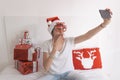 young beautiful woman sitting on bed and taking a selfie with mobile phone. Presents on bed. Wearing red santa hat and funny Royalty Free Stock Photo