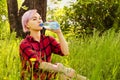 Young beautiful woman sitting in green grass, holds and drink bottle of water on a tree and bush background Royalty Free Stock Photo