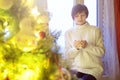 Young beautiful woman sitting home by the window with cup of hot coffee wearing knitted warm sweater. Christmas tree Royalty Free Stock Photo