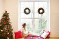 Young beautiful woman sitting home by the window wearing knitted warm sweater. Christmas tree with decorations and lights in the Royalty Free Stock Photo