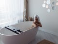 Young beautiful woman sitting in modern empty bathtub with towel on head in own apartment. Caucasian sexy female in bath Royalty Free Stock Photo