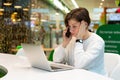 Young beautiful woman sitting at a shopping center at a table and working at a computer laptop, using mobile phone Royalty Free Stock Photo