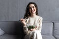 Young and beautiful woman sitting on sofa at home, eating colorful fresh vegetable salad and smiling, healthy woman enjoying food Royalty Free Stock Photo