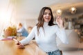 Young beautiful woman with smarphone in her hand waving her friend l while sitting in the cafe during lunch break, positive vibes