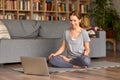 Young happy woman having online yoga class on laptop at home, sitting in lotus pose on floor