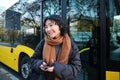 Young beautiful woman standing on bus stop, texting message on smartphone, holding mobile phone, checking her schedule