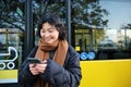 Young beautiful woman standing on bus stop, texting message on smartphone, holding mobile phone, checking her schedule