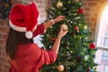 Young beautiful woman standing with happy face decoring christmas tree with glass baubles ornaments and wearing santa claus hat