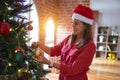 Young beautiful woman standing with happy face decoring christmas tree with glass baubles ornaments and wearing santa claus hat