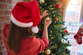 Young beautiful woman standing with happy face decoring christmas tree with glass baubles ornaments and wearing santa claus hat