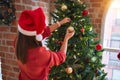 Young beautiful woman standing with happy face decoring christmas tree with glass baubles ornaments and wearing santa claus hat