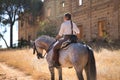 Young and beautiful woman standing, riding her horse on a country road next to an abandoned and ruined building, under the rays of