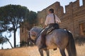 Young and beautiful woman standing, riding her horse on a country road next to an abandoned and ruined building, under the rays of