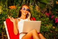 Young beautiful woman traveler freelancer sitting on a sun lounger with laptop talking on the phone on the seashore Royalty Free Stock Photo