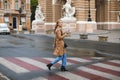 Young beautiful woman in trench coat and jeans thoughtfully looking in camera holding cellphone in hand while walking on