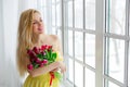 Young beautiful woman with tulip bunch in yellow dress looks at window. Royalty Free Stock Photo