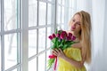 Young beautiful woman with tulip bunch in yellow dress looks at window. Royalty Free Stock Photo