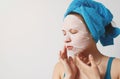 A young beautiful woman uses a moisturizing cosmetic fabric face mask with a towel wrapped around her head. on white background Royalty Free Stock Photo