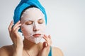 A young beautiful woman uses a moisturizing cosmetic fabric face mask with a towel wrapped around her head. on white background Royalty Free Stock Photo