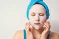 A young beautiful woman uses a moisturizing cosmetic fabric face mask with a towel wrapped around her head. on white background Royalty Free Stock Photo