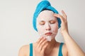 A young beautiful woman uses a moisturizing cosmetic fabric face mask with a towel wrapped around her head. on white background Royalty Free Stock Photo