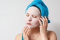 A young beautiful woman uses a moisturizing cosmetic fabric face mask with a towel wrapped around her head. on white background Royalty Free Stock Photo
