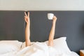 Young beautiful woman waking up in the morning in the bed, hiding under the blanket, stretching out her arms with a cup of coffee Royalty Free Stock Photo