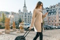 Young beautiful woman walks along city street with travel suitcase and cell phone. Fashionable brunette girl smiling Royalty Free Stock Photo