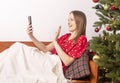 Young beautiful woman waving hand and saying hello while chatting by video call on mobile phone, lying on sofa in pygamas, wrapped