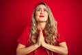Young beautiful woman wearing basic t-shirt standing over red isolated background begging and praying with hands together with Royalty Free Stock Photo