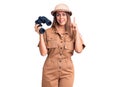 Young beautiful woman wearing explorer hat holding binoculars surprised with an idea or question pointing finger with happy face,