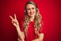 Young beautiful woman wearing headphones over red isolated background smiling with happy face winking at the camera doing victory Royalty Free Stock Photo