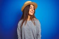 Young beautiful woman wearing navy striped t-shirt and hat over isolated blue background looking away to side with smile on face, Royalty Free Stock Photo