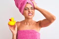 Young beautiful woman wearing shower towel holding duck over isolated white background stressed with hand on head, shocked with Royalty Free Stock Photo