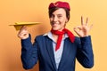Young beautiful woman wearing stewardess uniform holding paper plane doing ok sign with fingers, smiling friendly gesturing
