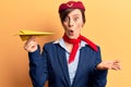 Young beautiful woman wearing stewardess uniform holding paper plane scared and amazed with open mouth for surprise, disbelief