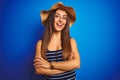 Young beautiful woman wearing striped t-shirt and summer hat over isolated blue background happy face smiling with crossed arms Royalty Free Stock Photo