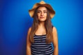 Young beautiful woman wearing striped t-shirt and summer hat over isolated blue background with serious expression on face Royalty Free Stock Photo