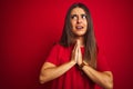 Young beautiful woman wearing t-shirt standing over isolated red background begging and praying with hands together with hope Royalty Free Stock Photo