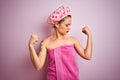 Young beautiful woman wearing towel and bath hat after shower over pink isolated background showing arms muscles smiling proud Royalty Free Stock Photo