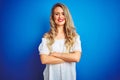 Young beautiful woman wearing white dress standing over blue isolated background happy face smiling with crossed arms looking at Royalty Free Stock Photo