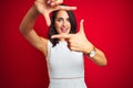 Young beautiful woman wearing white dress standing over red isolated background smiling making frame with hands and fingers with Royalty Free Stock Photo