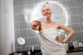 Young beautiful woman wearing white towel holding pink gouache scraper for face massage while doing beauty morning routine in Royalty Free Stock Photo