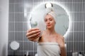 Young beautiful woman wearing white towel holding pink gouache scraper for face massage while doing beauty morning routine in Royalty Free Stock Photo