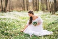 Young beautiful woman in a white dress collects primroses. A girl in the spring forest. A bouquet of white anemones in her hands