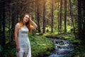 Young beautiful woman in white dress near icy stream on evergreen trees background in natural sunlight. Romantic nymph in summer Royalty Free Stock Photo