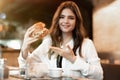 Young beautiful woman in white stylish blouse looks hungry pointing with her hand at fresh meat burger during lunch in trendy cafe Royalty Free Stock Photo