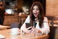 Young beautiful woman in white stylish jacket with smartphone in her hands looking shocked during coffee break in cafe modern Royalty Free Stock Photo