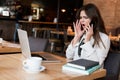 Young beautiful woman working in her laptop looking angry during phone conversation while drinking hot coffee in the cafe Royalty Free Stock Photo